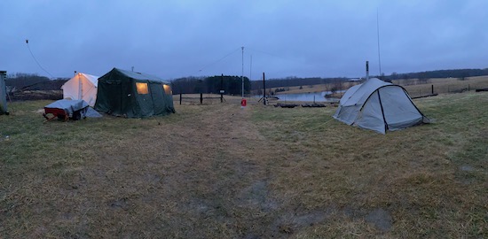 A rain-soaked field with 3 large hot-tents, various antennas and a pond in the background